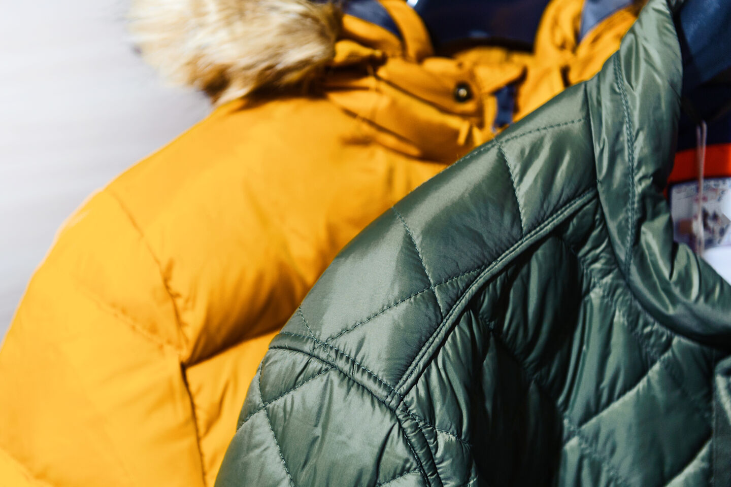 A close-up of two down jackets. The yellow one has a fur lining on the hood, while the green one does not. A close-up of two down jackets. The yellow one has a fur lining on the hood, while the green one does not.