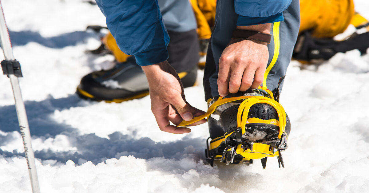 A close-up of a man bending over, putting his snow boots into a pair of yellow, rigid crampons.