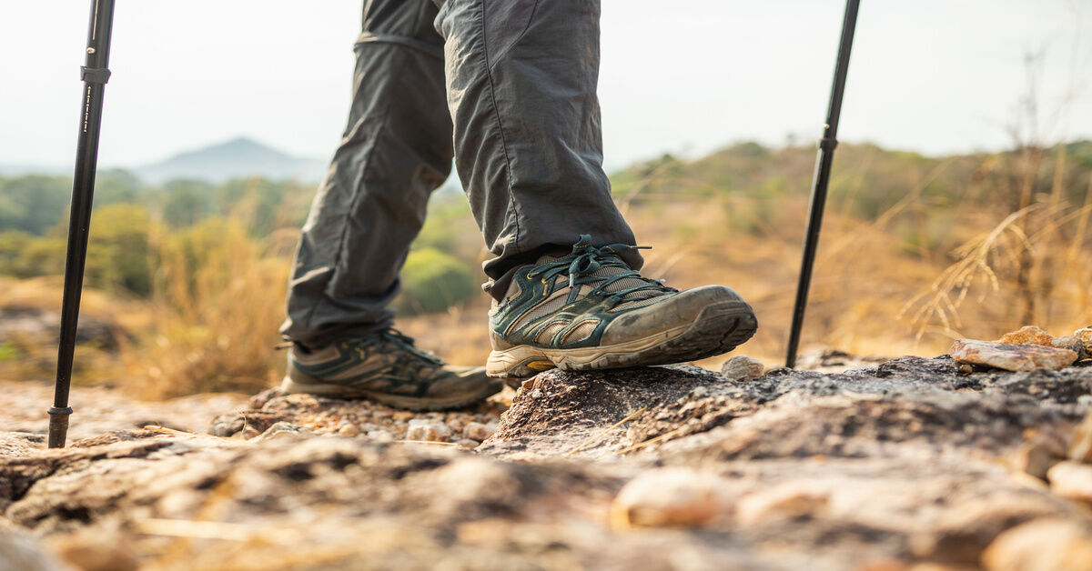 A close-up of a person wearing mountain boots and using trekking poles to cross a rocky terrain.